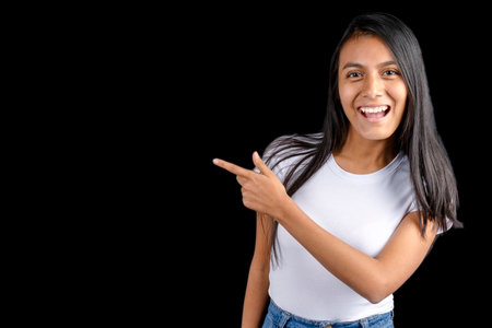 Beautiful Latin woman on a black background pointing with her index finger and smiling, looking at the indicated space.の写真素材