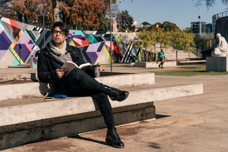Man sitting on a concrete bench in a public park reading a book and having a hot drink in the sun.