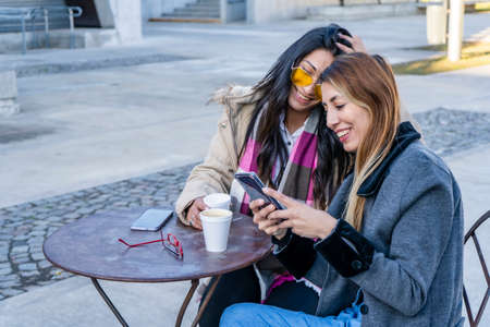 Two happy women latin friends or sisters sharing a smart phone in a coffee shop terrace looking at device in winter.の写真素材