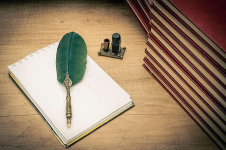 Old books stacked in disarray, an old pen with its ink and a notepad on an old wooden table and dim lighting. Selective focus. Vintage scene concept.の写真素材