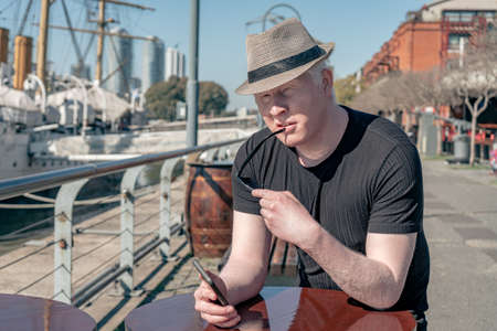 Medium shot of an albino man in a hat checking his cell phone messages while sitting in a bar in the port.の写真素材