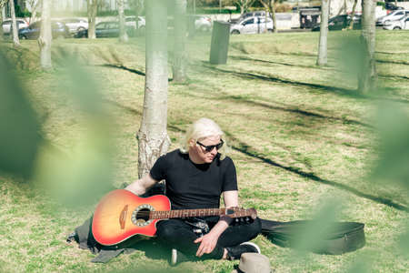 Young albino man sitting on the grass leaning against a tree relaxed with his guitar resting on his...