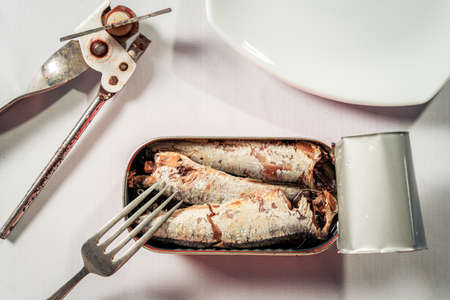 Open can of sardines in oil next to a can opener, a plate and a fork on a white marble-like surface. Marine food productの写真素材