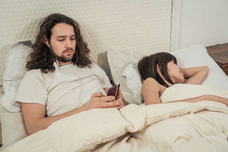 Couple in bed. The man listening to music with headphones while the woman sleeps. Concept rest, love, respect.の写真素材