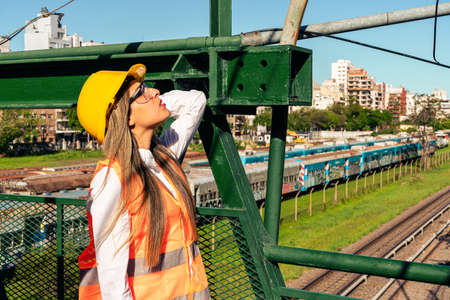 Beautiful young female engineer wearing safety equipment, inspects a railroad pedestrian bridge.