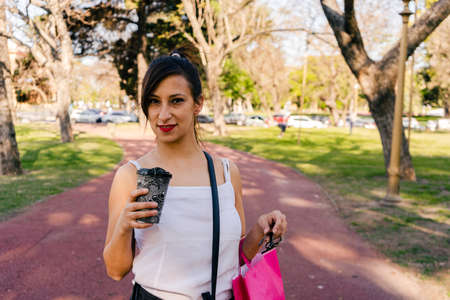A Portrait of a beautiful Latin woman walking through a park with shopping bags and drinking a coffee. Shopping concept.の写真素材