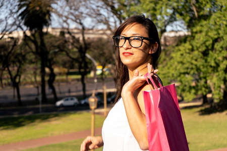 Portrait of a beautiful Latin woman with glasses and casual clothes walking happily through a park with shopping bags. Shopping concept.の写真素材