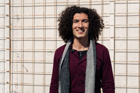 Short medium shot Portrait of a smiling young Latino in jean and burgundy shirt, with curly hair,...