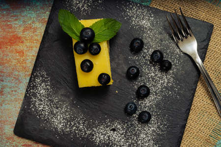 One Portion of homemade lemon curd cake in the shape of an ingot or brick decorated with some mint leaves and blueberries on a black slab. Top view. Copy space.の写真素材