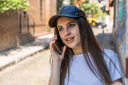 A portrait of a beautiful teenage girl smiling and wearing a black cap talking with cell phone or...