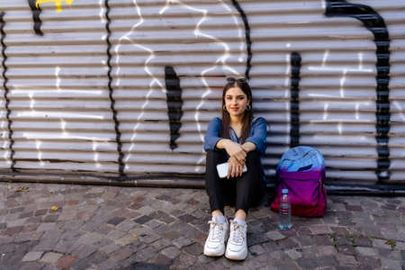 A Beautiful young Hispanic woman sitting on the street resting smiling with her backpack at her side and her cell phone in her hand.の写真素材