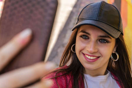 Close up portrait of a young attractive woman holding a smartphone digital camera with her hands and taking a selfie self portrait of herself winking at he camera. Travel and technology outdoors.の写真素材