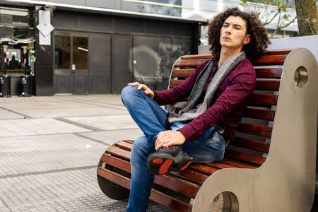 A Young curly haired man sitting on a bench in the street and waiting with serious expression.