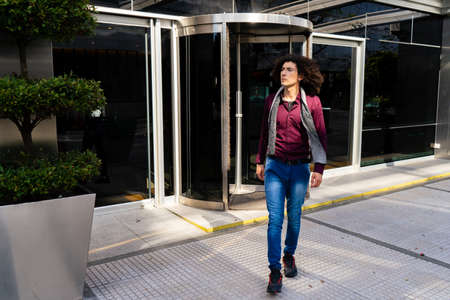 A Young curly haired Latino man walking out of a revolving door building with a worried or serious expression.の写真素材