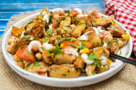 A delicious potato, chickpea, tomato and mushroom salad with parsley and aioli in a plate on a wooden table. Healthy, homemade, vegan food. Low view.の写真素材