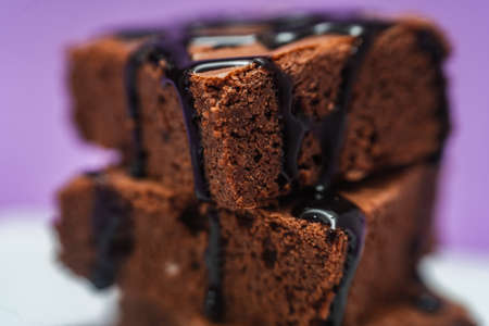Very close shot of a Pile of Chocolate Brownies with chocolate strands falling on a white plate with purple background.の写真素材