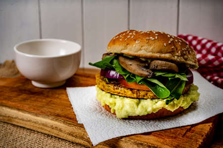 Vegan veggie protein burger, with tomato, arugula, red onion, mushrooms and avocado mayonnaise on a wooden board and a white background. Chopped view. Copy text. Natural food concept, vegetarian.の写真素材