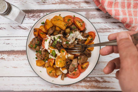 Hand taking a portion of beef chunks sauteed with cassava or manioc, chives, pepper, lemon and cherry tomato on a rustic surface. top viewの写真素材