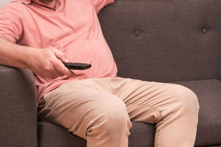 unrecognizable man sitting on a sofa with a remote control in his hand changing channels on the...