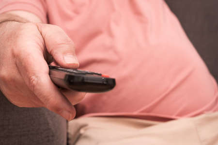 unrecognizable man sitting on a sofa with a remote control in his hand changing channels on the television. concept of rest and relaxationの写真素材