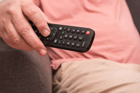 unrecognizable man sitting on a sofa with a remote control in his hand changing channels on the...