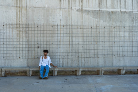 A young man with a personal style sitting on a bench in front of a large concrete wall.
