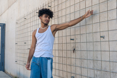 A handsome young Latino man with a personal style posing and holding a fence of a large concrete wall.の写真素材