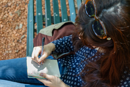 Writer taking notes in spiral notebook while sitting on park bench. Focus on Hands