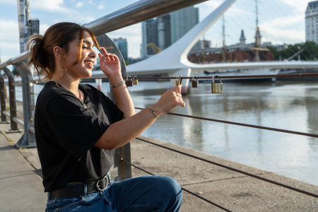 Happy young woman placing a love lock on the wire fence along Puerto Madero waterfront with the...