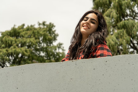 Portrait of a cheerful young woman with long dark hair and wearing a red and black checkered shirt,...