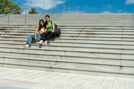 Two young women with backpacks are sitting on urban steps, enjoying a break in the sunshineの写真素材