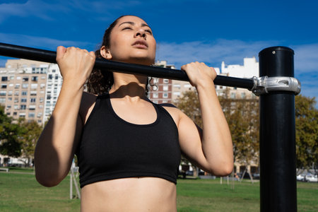 Determined athletic woman exercising outdoors, performing pull-ups on a horizontal bar, promoting...