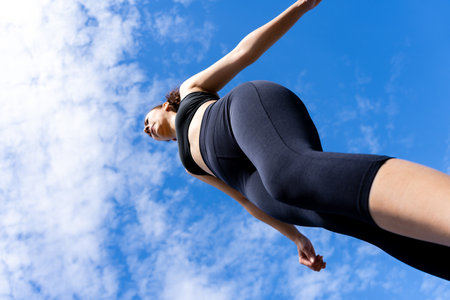 Low angle view of sporty young woman walking under blue cloudy sky, wearing black sportswear