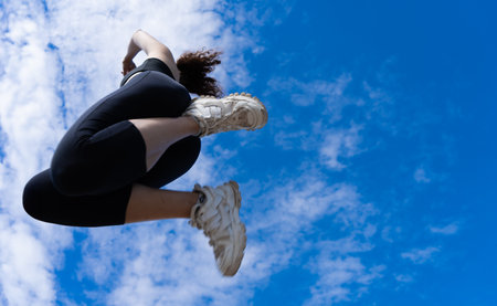 Sportswoman performing a powerful jump against a backdrop of blue sky and clouds, showcasing athleticism and determinationの写真素材
