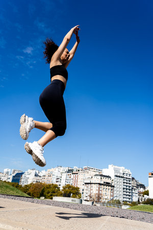 Female athlete performing a jump with arms raised, showcasing athleticism and urban fitness against...