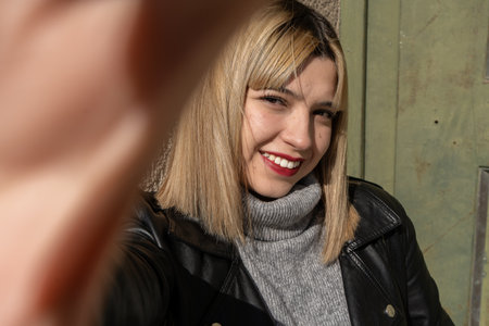 Happy young woman taking selfie and smiling while wearing leather jacket and turtleneck sweater