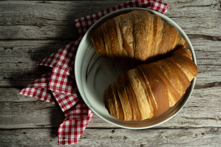 Two golden croissants on a porcelain plate with a gingham napkin atop a wooden table. Perfect for food blogs or lifestyle stock images showcasing warmth and simplicity in culinary presentations.の写真素材