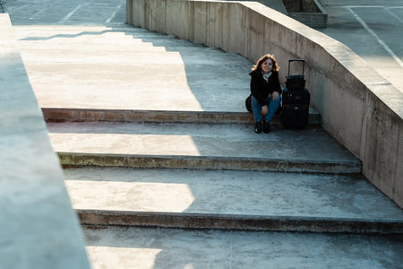 Young woman resting seated on wide concrete stairs with a suitcase by her side, waiting patiently.の写真素材