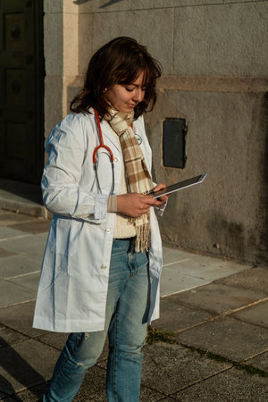 Young female doctor in a white coat walks along the sidewalk in front of a medical center while...