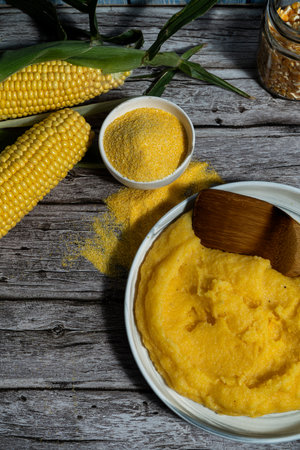 Creamy polenta served on a plate, accompanied by corncobs, corn flour, and a wooden spoon, creating a rustic culinary sceneの写真素材