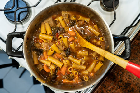 Delicious beef stew with pasta and vegetables cooking in a pot on the stove, being stirred with a...