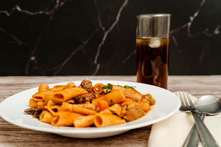 Steaming hot pasta dish with beef, vegetables, and tomato sauce, served with a refreshing drink on a wooden tableの写真素材