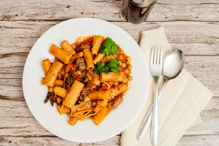 Pasta with meat and vegetables, also known as beef stew, is served on a white plate on a rustic wooden table with cutlery and a bottle of drinkの写真素材