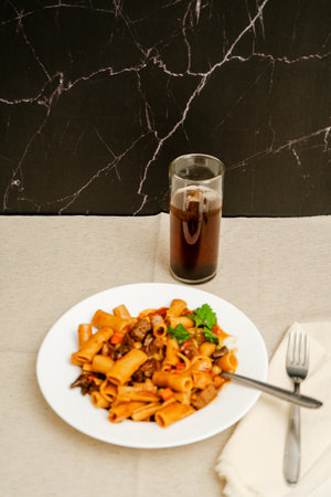 Savory pasta dish with meat and vegetables, served alongside a dark beverage, creating a satisfying...
