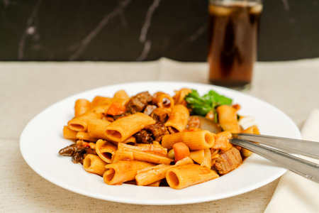 Steaming hot rigatoni pasta with beef, vegetables, and fresh basil, served alongside a refreshing glass of colaの写真素材