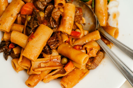 Rigatoni pasta mixed with beef stew, carrots, peas and tomato sauce being mixed with two silver cutlery pieces on a white plateの写真素材