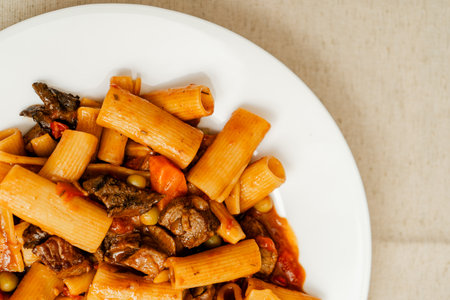 Beef stew with rigatoni pasta, peas, carrots, and mushrooms served on a white plate, offering a hearty and flavorful mealの写真素材