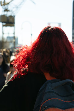 Woman with vibrant, flowing red hair walks down a bustling city street, her backpack slung over her shoulderの写真素材