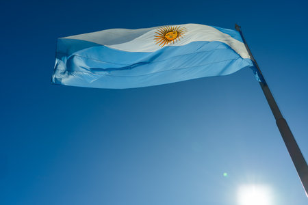 The Argentinian flag waving proudly in the wind against a clear blue sky, illuminated by the sunの写真素材