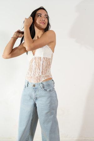 Fashionable young latin woman wearing a white lace bodysuit and light blue jeans, touching her long dark hair, smiling and looking awayの写真素材